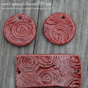 May include: Three red clay pottery pendants with a swirl design. The pendants are round and rectangular in shape. The pendants have holes for hanging.