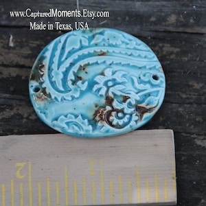 May include: A turquoise blue ceramic oval pendant with a paisley design. The pendant has a rustic, hand-crafted look with brown accents.