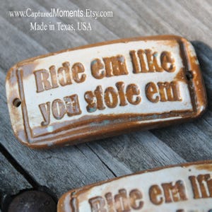 May include: A brown and white ceramic sign with the text "Ride em like you stole em" in brown lettering. The sign has a rustic, distressed look.