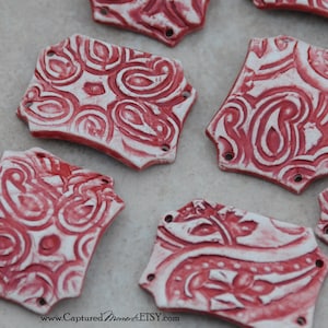 May include: Six red and white ceramic tiles with a paisley pattern. The tiles are rectangular and have rounded corners. They have holes in the corners for hanging.