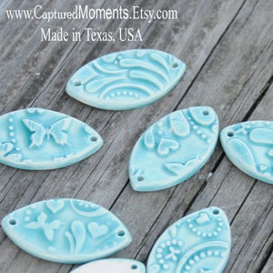 May include: Six aqua blue ceramic oval-shaped pendants with various embossed designs, including butterflies, hearts, and swirls. The pendants have two holes for hanging.