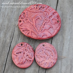 May include: Three red ceramic pendants with a floral design. The largest pendant is oval shaped and the two smaller pendants are round. All three pendants have a hole for stringing.