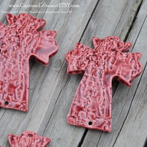 May include: Two red ceramic cross pendants with a textured surface. The crosses are handmade and have a hole at the top for hanging.