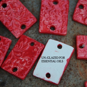 May include: A collection of red ceramic tiles with a floral design and two holes. The tiles are unglazed and are labeled "UN-GLAZED FOR ESSENTIAL OILS".