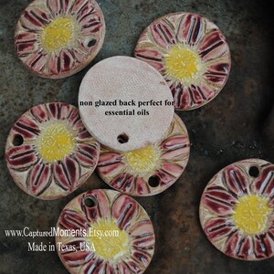 May include: Handmade ceramic flower-shaped beads with a yellow center and red-burgundy petals. The beads have a hole for stringing and a non-glazed back. The text on the back says "non glazed back perfect for essential oils".