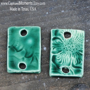 May include: Two green ceramic tiles with holes for hanging. One tile has a floral design and the other has a swirling pattern.