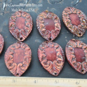 May include: Six ornate, oval-shaped metal charms with a copper finish and intricate scrollwork designs. The charms have a reddish-brown patina and a hole at the top for hanging.