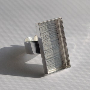 May include: A silver-toned adjustable ring with a rectangular bezel. The bezel has a textured surface, and the ring is set against a white background, casting a shadow.