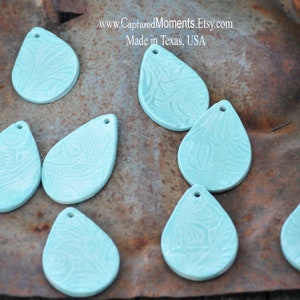 May include: Ten teal blue ceramic teardrop-shaped pendants with a decorative embossed pattern. The pendants have a hole at the top for hanging.