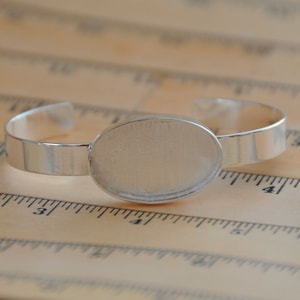 May include: Silver bracelet with a round, blank, silver-toned bezel. The bracelet is lying on a wooden ruler.