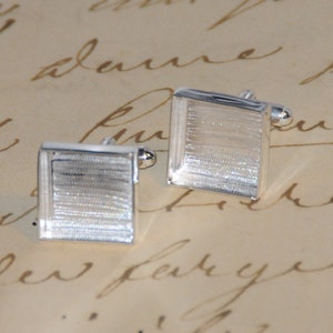 May include: A pair of silver cufflinks with a textured, rectangular design. The cufflinks have a shiny finish and are set against a cream-colored background.