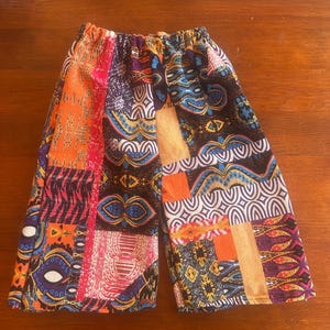 Puede incluir: Pantalones cortos de patchwork coloridos con cintura elástica. Los pantalones cortos presentan una variedad de patrones en naranja, rojo, azul y negro. La tela parece ser un material ligero, adecuado para climas cálidos.