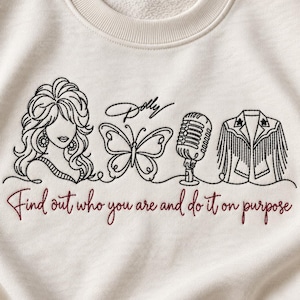 May include: A cream-colored crewneck sweatshirt featuring black line-art embroidery of a woman, a butterfly, a microphone, and a fringed jacket. The text "Dolly" and "Find out who you are and do it on purpose" is embroidered in black and red.