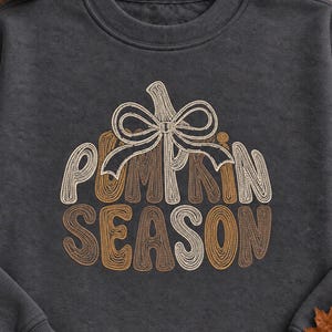 Puede incluir: Una sudadera de cuello redondo gris carbón con un gráfico bordado en forma de calabaza formado por el texto PUMPKIN SEASON en tonos crema, bronceado y marrón, rematado con un lazo decorativo bordado en un hilo de color crema a juego.