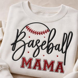 Pode incluir: Uma camisola de gola redonda cor de creme com texto bordado que diz Baseball MAMA. A palavra Baseball está em cursivo preto com um gráfico de basebol vermelho e preto por cima, e MAMA está em letras de bloco vermelhas abaixo.