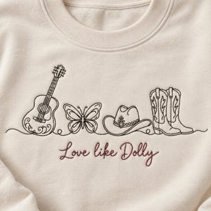 Puede incluir: Una sudadera de cuello redondo color crema con bordado de líneas negras que representa una guitarra acústica, una mariposa, un sombrero de vaquero y un par de botas de vaquero. Debajo de las ilustraciones, el texto "Love like Dolly" está bordado en rojo oscuro.