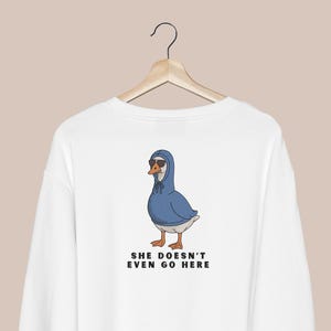 Peut inclure: Sweat-shirt blanc avec un dessin d'oie portant des lunettes de soleil et un sweat &agrave; capuche bleu. L'oie a des pieds orange et un bec jaune. Le texte "SHE DOESN'T EVEN GO HERE" est imprim&eacute; en dessous.