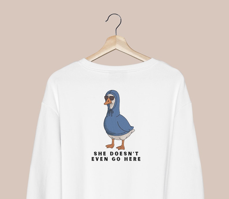 Peut inclure: Sweat-shirt blanc avec un dessin d'oie portant des lunettes de soleil et un sweat &agrave; capuche bleu. L'oie a des pieds orange et un bec jaune. Le texte "SHE DOESN'T EVEN GO HERE" est imprim&eacute; en dessous.