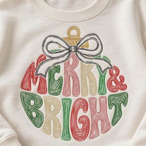 May include: A cream-colored crewneck sweatshirt featuring an embroidered Christmas ornament design. The ornament is filled with the text MERRY & BRIGHT in red, green, and gold thread, topped with a large white bow with black outlines.