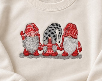 May include: A cream-colored crewneck sweatshirt featuring an embroidered design of three gnomes standing in a row. The two outer gnomes wear red hats with white patterns and have long white beards. The center gnome wears a black and white plaid hat.