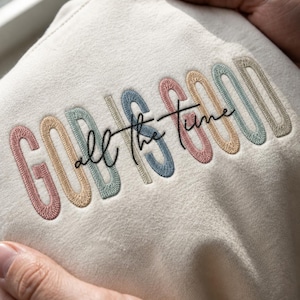 Può includere: Una felpa color crema con il testo ricamato GOD IS GOOD in fili alternati rosa pastello, beige, verde salvia e blu. Le parole all the time sono scritte in un carattere corsivo nero al centro del ricamo sul tessuto della felpa.