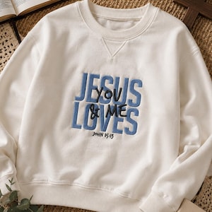 May include: An off-white crewneck sweatshirt featuring blue embroidered text that reads JESUS LOVES YOU & ME, with the smaller black embroidered text JOHN 15:13 positioned directly underneath the main phrase on the front of the garment.