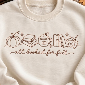 Puede incluir: Una sudadera de cuello redondo color crema con un bordado de líneas marrones que muestra una calabaza, libros apilados, una taza con crema batida, más libros y una hoja de arce, junto al texto "all booked for fall" en letra cursiva.