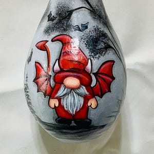 May include: A hand-painted gourd ornament featuring a red gnome with devil wings and a white beard. The gnome is standing on a gray background with black bats.