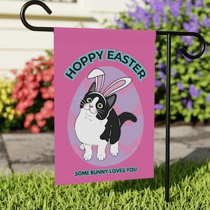 Puede incluir: Una bandera de jardín rosa con un gato blanco y negro con orejas de conejo. La bandera dice "Hoppy Easter" y "Some Bunny Loves You".