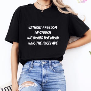 Puede incluir: Una camiseta negra de manga corta con texto en blanco que dice: WITHOUT FREEDOM OF SPEECH WE WOULD NOT KNOW WHO THE IDIOTS ARE. La camiseta tiene cuello redondo y se combina con pantalones cortos de mezclilla azul con detalles desgastados.