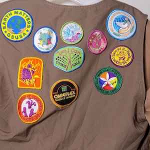 Könnte beinhalten: Eine braune Weste mit mehreren bunten Aufnähern. Auf den Aufnähern stehen Texte wie "Earth Matters", "Girl Scouts", "Yes I Can Cookie Sale" und "Omniplex Science Museum". Die Weste hat ein hellbraunes Futter.