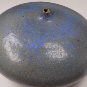 Puede incluir: Un jarrón redondo de cerámica con un esmalte gris y azul. El jarrón tiene una pequeña abertura en la parte superior y una forma lisa y redondeada. El esmalte azul se concentra en el centro, creando un contraste llamativo con el gris.