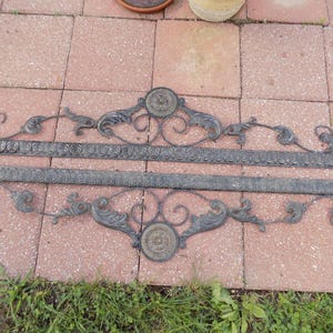 Può includere: Due elementi architettonici allungati in metallo scuro con un medaglione circolare centrale a motivo floreale. Ogni pezzo presenta un bordo lineare con un motivo a foglie ripetuto e volute decorative che si estendono dal centro.