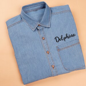 Könnte beinhalten: Ein hellblaues Jeanshemd mit klassischem Kragen und einer einzelnen Brusttasche. Das Hemd hat braune Knöpfe und Nähte. Der Name "Delphine" ist in schwarzer Schrift auf der Tasche gestickt.