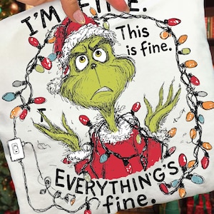 May include: White t-shirt featuring a Grinch character wearing a Santa hat and Christmas lights. The text reads "I'm fine. This is fine. Everything's fine." The Grinch has a green face and is wearing a red Santa suit.