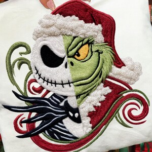 May include: White Christmas-themed pillow featuring a split design of Jack Skellington and the Grinch wearing Santa hats. The embroidery includes green, red, black, and white details, with swirling accents. A festive holiday decoration.
