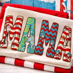 Puede incluir: Una camiseta blanca con la palabra "MAMA" en letras grandes y coloridas. Las letras presentan diseños inspirados en los personajes y patrones del Dr. Seuss, incluyendo rayas rojas y blancas, zigzags azules e ilustraciones caprichosas. La camiseta está doblada sobre tela a rayas rojas y blancas.