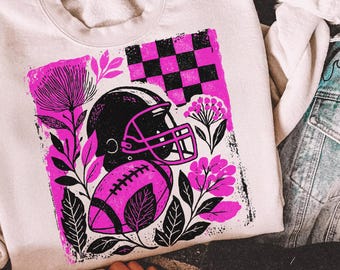 May include: Cream-colored sweatshirt featuring a graphic of a black football helmet and pink football, with pink floral accents and a checkered flag design. The sweatshirt is paired with a denim jacket and checkered slip-on shoes.