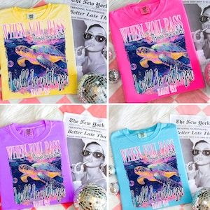 May include: Four colorful t-shirts: yellow, pink, purple, and blue. Each shirt features the text "WHEN YOU PASS Through deep waters I will be with you" with a sea turtle graphic. A newspaper and disco ball are in the background.