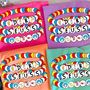 May include: Four colorful t-shirts, each featuring a Dr. Seuss-themed design. The design includes the text "DOR SEUSS" in a bracelet-style graphic with various Dr. Seuss character illustrations. The shirts are in pink, purple, orange, and teal.
