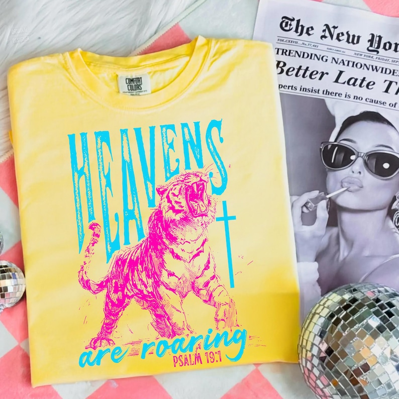 May include: Yellow t-shirt with a graphic of a roaring pink tiger and the text "HEAVENS ARE ROARING" in blue. The shirt also features the text "PSALM 19:1". A newspaper and disco ball are in the background.