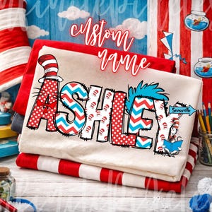 May include: A stack of folded t-shirts with the name "ASHLEY" in a whimsical, colorful design. The letters feature red polka dots, blue zigzags, and a Cat in the Hat theme. The words "Custom Name" are written above the name.