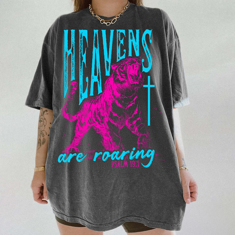 May include: Dark gray t-shirt with a vibrant pink tiger graphic and the words "HEAVENS are roaring" in blue. A cross is next to the tiger. The text "PSALM 19:1" is below the words "are roaring".