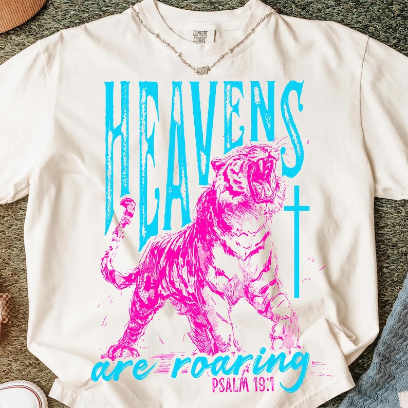 May include: Cream-colored t-shirt with a vibrant graphic. The design features the words "HEAVENS" in light blue, a roaring pink tiger, a light blue cross, and the words "are roaring" with "PSALM 19:1" below.