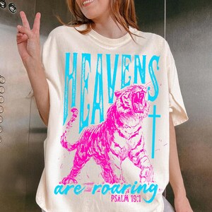 May include: Cream-colored t-shirt with a vibrant graphic. The design features a roaring pink tiger and the words "HEAVENS ARE ROARING" in blue, with "PSALM 19:1" below. A cross is also included in the design.
