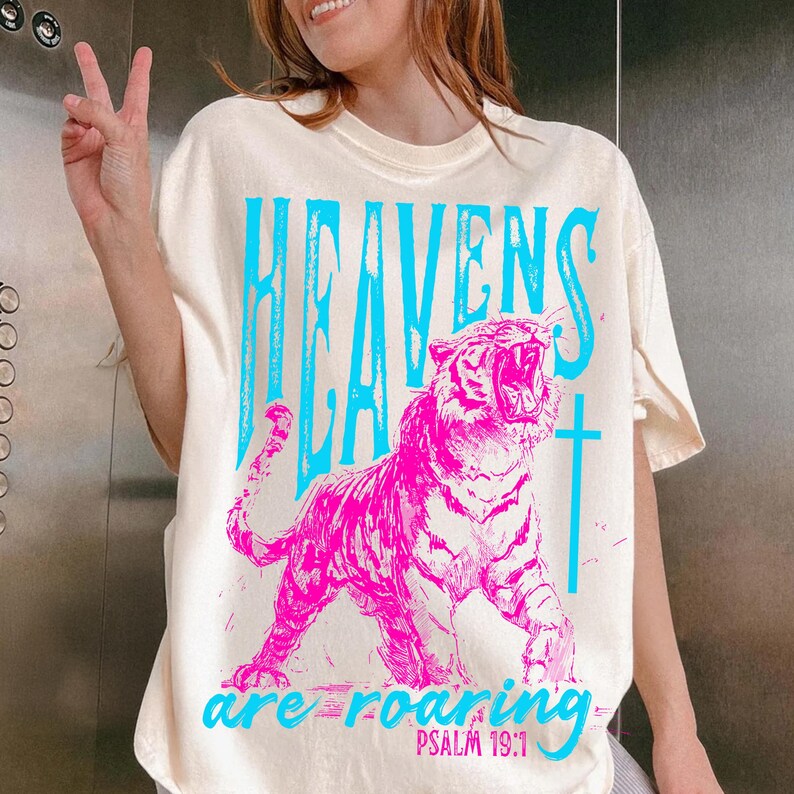May include: Cream-colored t-shirt with a vibrant graphic. The design features a roaring pink tiger and the words "HEAVENS ARE ROARING" in blue, with "PSALM 19:1" below. A cross is also included in the design.