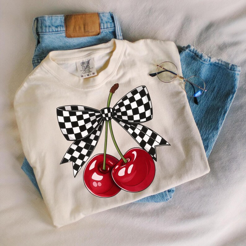 Puede incluir: Camiseta color crema con un gr&aacute;fico de dos cerezas rojas con tallos verdes y un lazo a cuadros blanco y negro. La camiseta est&aacute; sobre unos vaqueros azules, con unas gafas redondas.