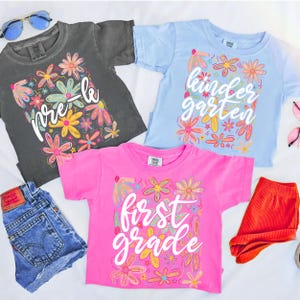 May include: Three colorful t-shirts with floral designs and school grade level text: "pre-k", "kindergarten", and "first grade". The shirts are gray, light blue, and pink. Also pictured are denim shorts and orange shorts.