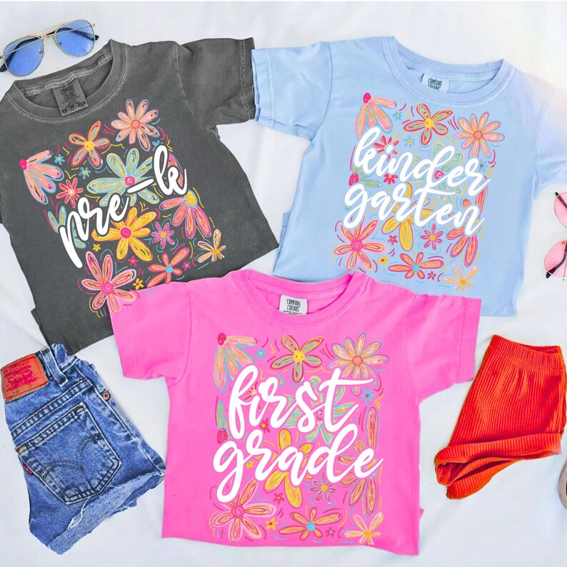 May include: Three colorful t-shirts with floral designs and school grade level text: "pre-k", "kindergarten", and "first grade". The shirts are gray, light blue, and pink. Also pictured are denim shorts and orange shorts.
