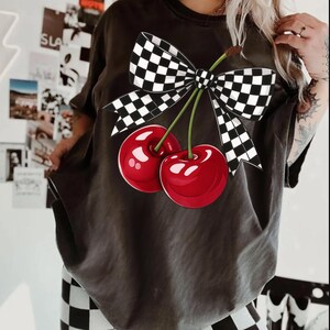 Puede incluir: Camiseta gris oscuro con un gr&aacute;fico de dos cerezas rojas brillantes, tallos verdes y un lazo a cuadros blanco y negro. La camiseta tiene un corte holgado y mangas cortas.